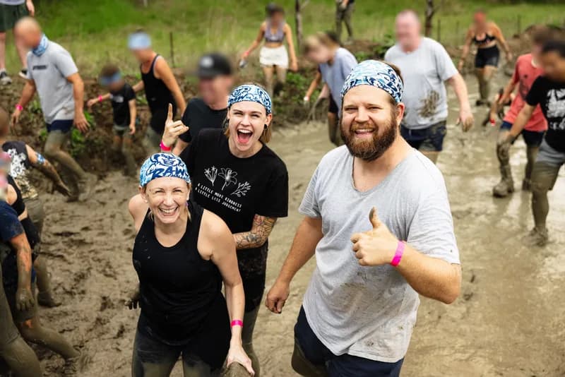 Three adults, wearing bandanas, standing in a pit of mud with other people, while smiling and looking at the viewer.