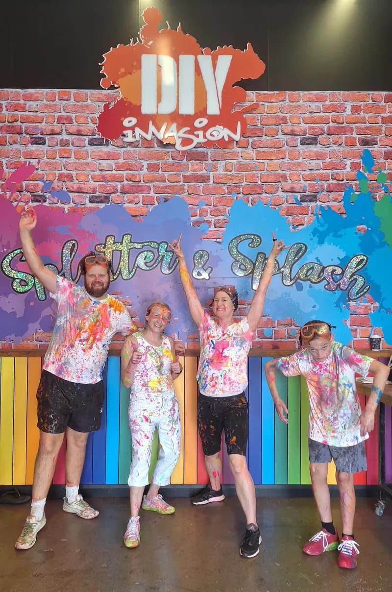 Three adults and a child, standing side-by-side, covered in colourful paint, looking at the viewer while smiling and cheering.