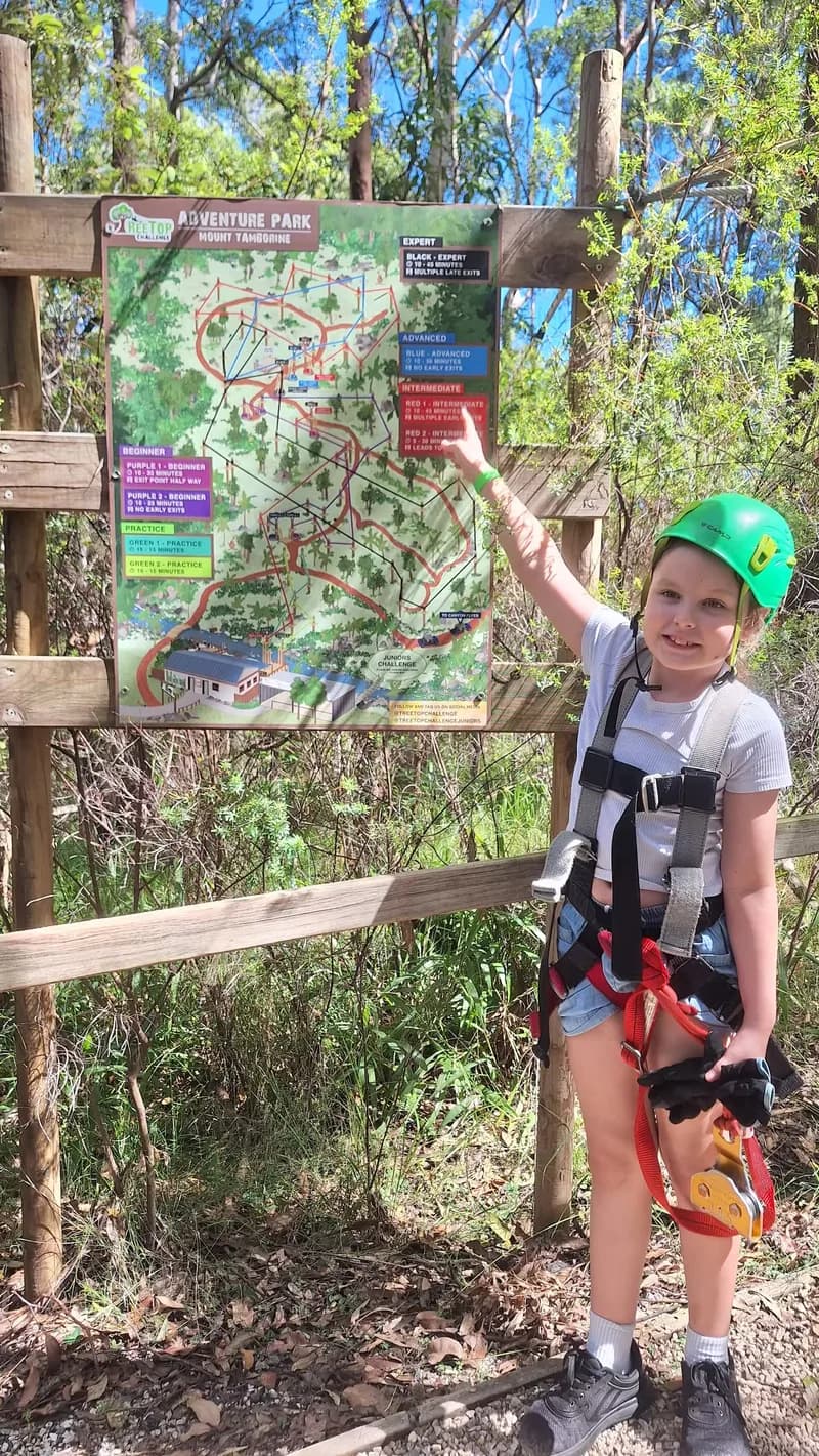 A child, wearing a helmet and safety harness, looking at the viewer, while pointing at a red-coloured section on a large high ropes course map.