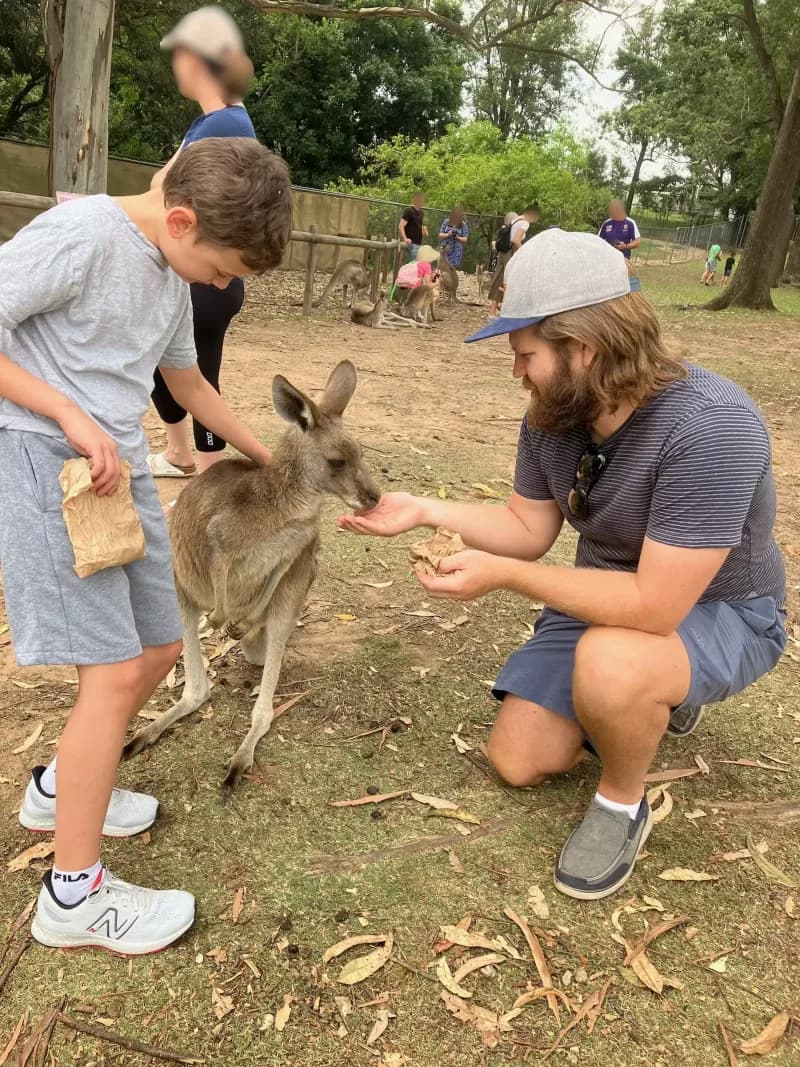 A therapist and child, interacting with a small kangaroo in an enclosure, the adult crouching on the grass feeding the kangaroo, the child patting it's back.