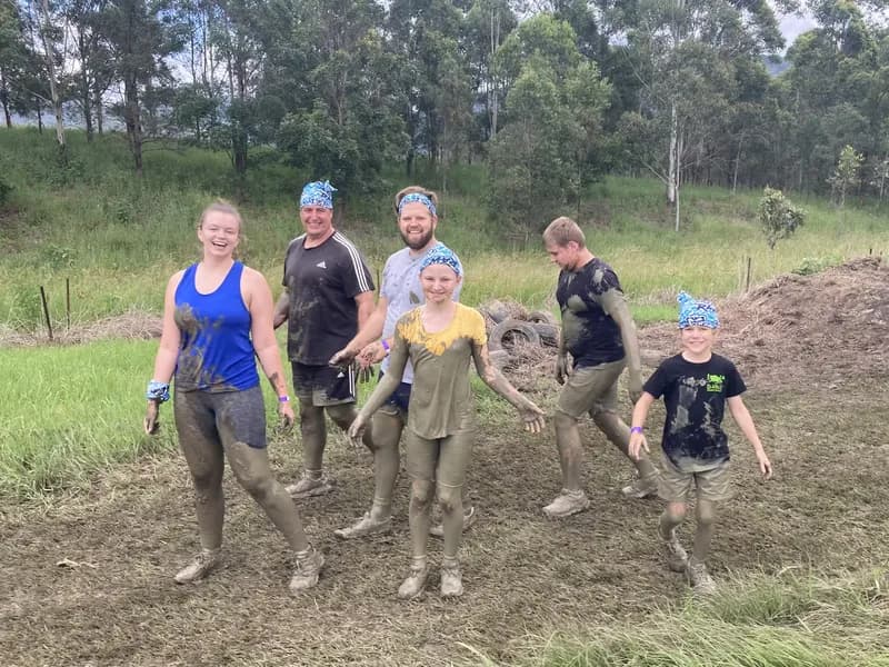 A group of 3 adults and 3 children, covered in mud and standing on a muddy track, looking at the viewer.