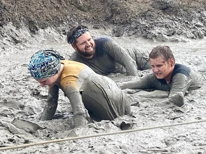 An occupational therapist and 2 children crawling through a muddy pit as part of an outdoor obstacle course.