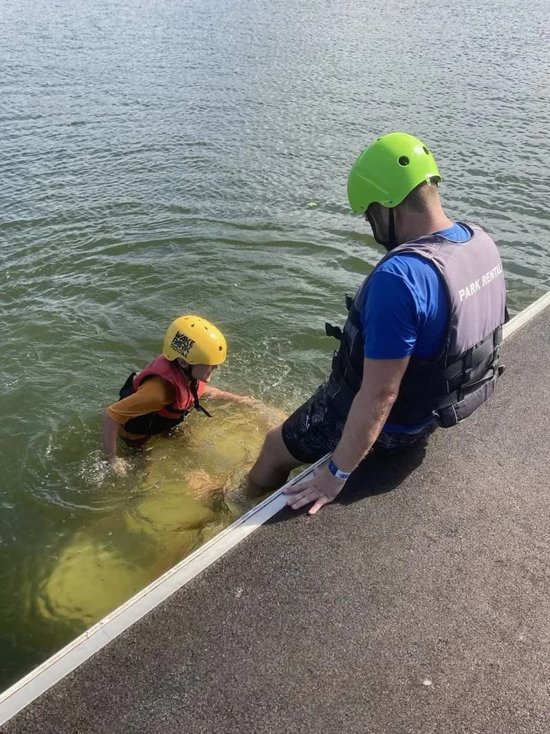 An occupational therapist helping a child balance on a water-ski board in the water.