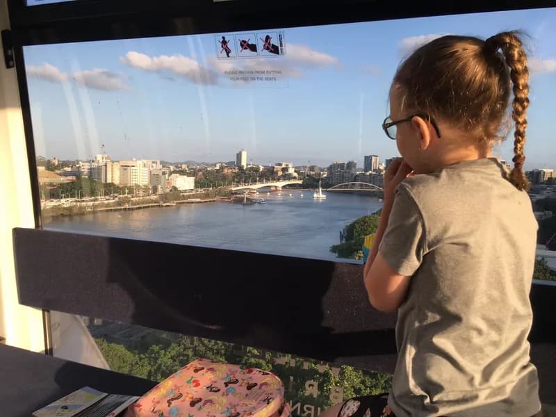 A child, sitting in an enclosed ferris wheel-style capsule high above the ground, looking out a glass window towards a river and city skyline.