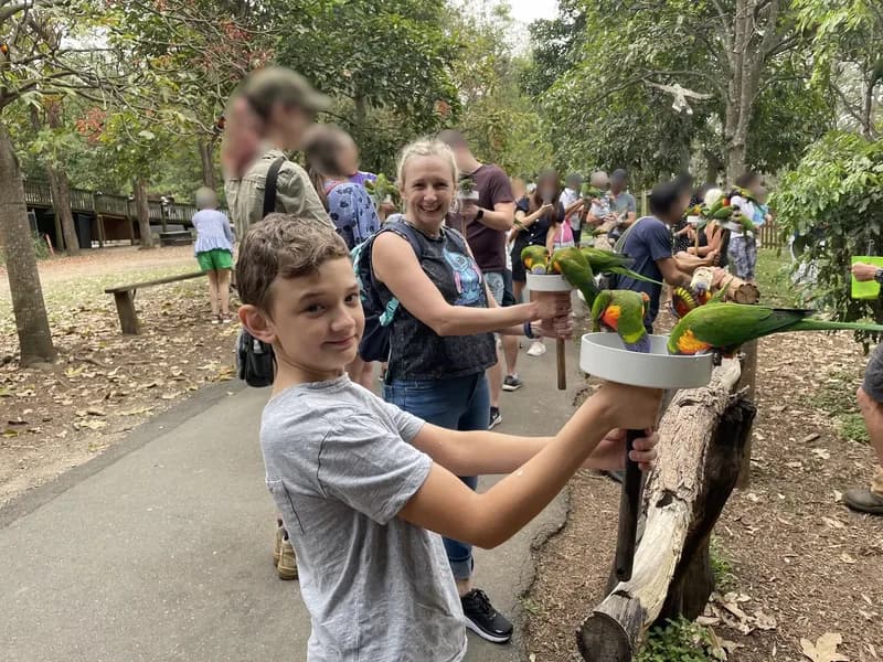 An adult and child, standing on an outdoor path among a group of people, looking at the viewer, holding bowls of bird food, with rainbow lorikeets sitting on the bowls eating the food.