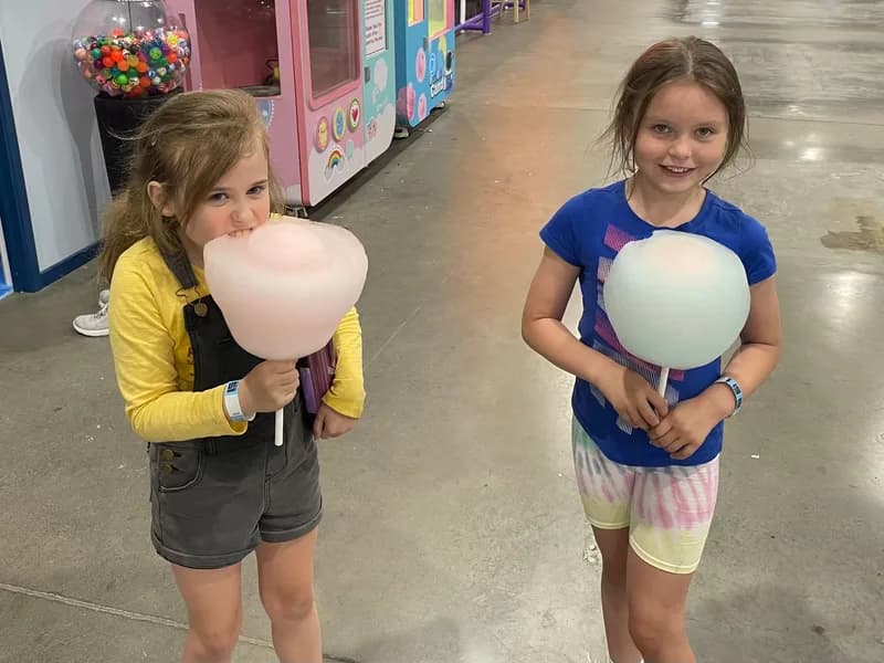 Two children, holding giant balls of fairy floss, one looking at the viewer, and one looking at and taking a bite of their fairy floss.