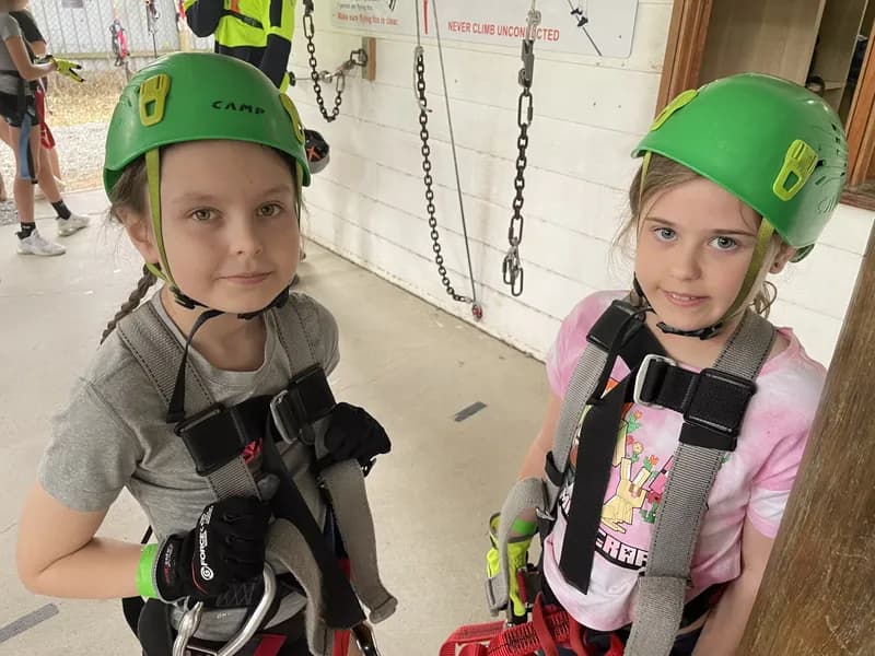 Two children, both wearing a helmet and safety harness, in front of climbing ropes and clips, appearing nervous and looking at the viewer.