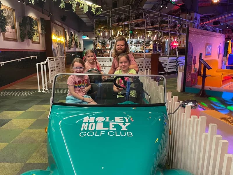 A therapist and 3 children, sitting in a novelty car beside a colourful indoor minigolf course, smiling and looking at the viewer.