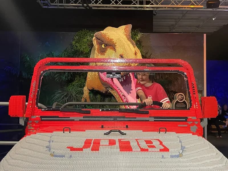A child, sitting in a life-size LEGO Jeep, looking in the rear-view mirror, with a large LEGO T-Rex chasing.