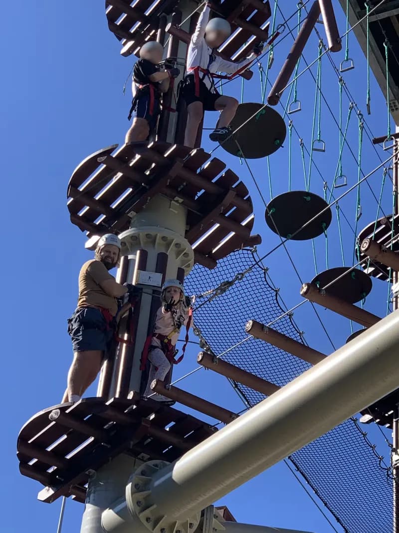 A therapist and child, wearing helmets and safety harnesses, standing on a platform high above the ground, about to complete a high ropes course, while looking at the viewer.