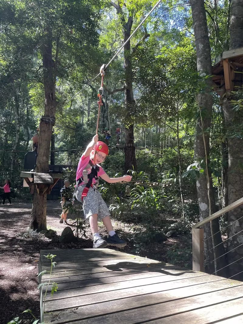 A child, wearing a helmet and safety harness, riding on a low zipline between trees, appearing excited and looking at a landing platform.