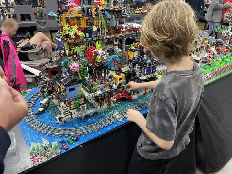 A child, standing at a table, while looking at and playing with a large LEGO display.