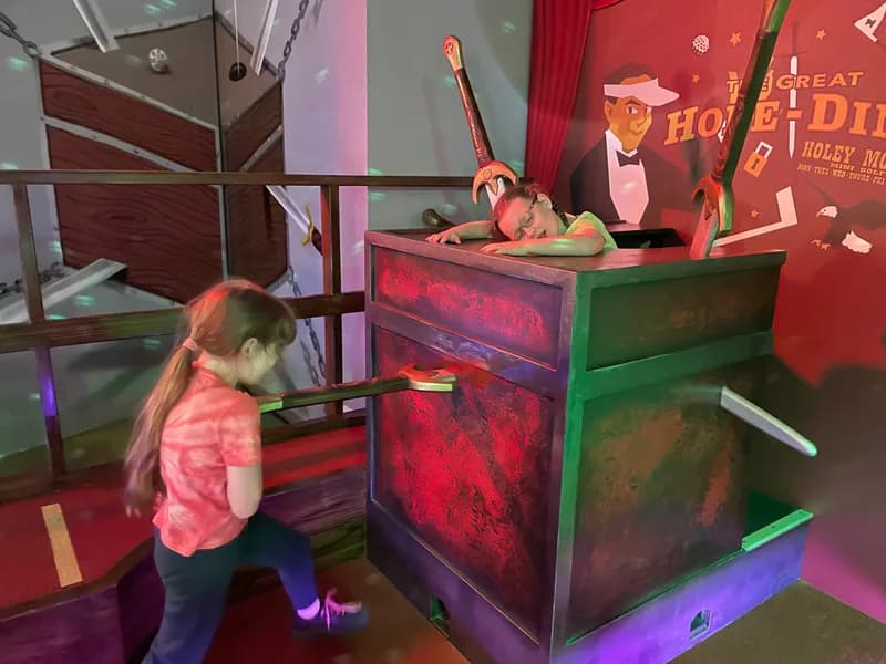 Two children, posing in an interactive scene, one pushing a sword into a box, the other standing in the box and pretending to be dead.
