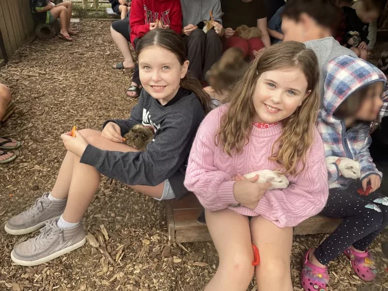 Two children, cuddling guinea pigs in their lap, sitting on a wooden log amongst a group of people, appearing happy and looking at the viewer.