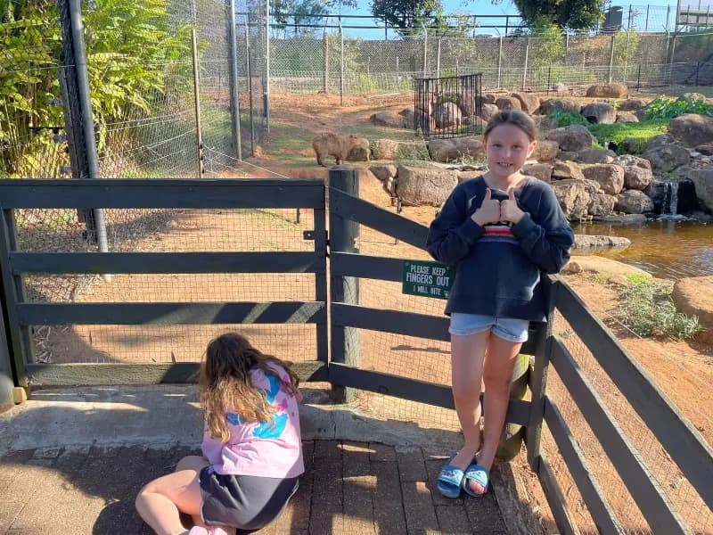 Two girls, in front of a capybara enclosure at a zoo, one standing and looking at the viewer giving a 'thumbs up', the other sitting and looking away from the viewer into the enclosure.