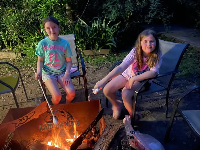 Two children, sitting in chairs around an outdoor firepit at night, toasting marshmallows, appearing happy while looking at the viewer.