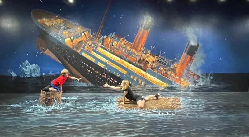 Two children, posing in an interactive art display of the sinking Titanic, one kneeling on a barrel while looking and reaching towards the other, who is laying on a piece of wooden debris and reaching towards the hand of the first child.