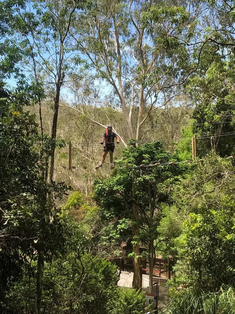 A therapist, wearing a helmet and safety equipment, standing on a rope bridge suspended between 2 large trees, looking at the viewer.